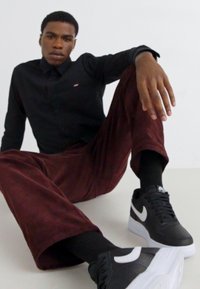 Črna srajca z gumbi, burgundske široke žametne hlače, črne Nike superge z belim logotipom, sedi na svetli površini v nevtralni ozadju.