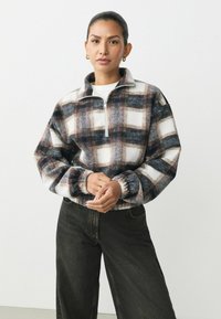 Fleece pullover i et mørkt ternet mønster med hvide accenter, der har en høj krave, halvlukket lynlås og samlede manchetter. Parret med mørk denim bukser.