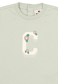 Camiseta de algodón verde claro con una gran letra 'C' estilizada en blanco, adornada con flores rosas y hojas verdes como detalles decorativos.