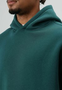 Groene hoodie van zacht stof, met een ruime capuchon, een ontspannen pasvorm en minimale naden. De textuur oogt glad.