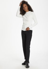 Haut à manches longues blanc à côtes avec un col et un ourlet à volants, associé à un pantalon noir sur mesure et des chaussures noires brillantes.