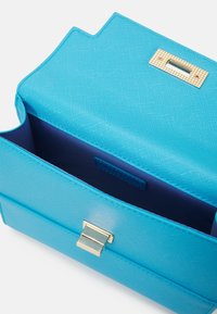 Turquoise handbag made of textured material. Features a gold clasp closure and a solid navy interior. Distinctive silhouette with sharp angles.