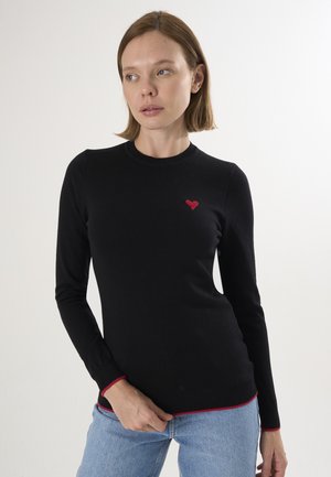 Donna che indossa un maglione nero a maniche lunghe con un piccolo cuore rosso sul petto sinistro e rifiniture rosse su polsini e orlo, abbinato a jeans azzurri chiari.