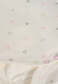 Crèmefärgad overall gjord av mjukt tyg, med små rosa och gröna blommönster. Den har rosa tryckknappar för stängning.