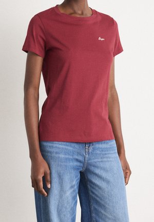 Vrouw draagt een nauwsluitend rood T-shirt met korte mouwen, voorzien van een klein wit logo op de borst, en lichtblauwe spijkerbroek tegen een effen achtergrond.