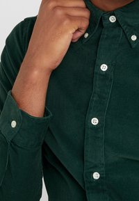 Zielona koszula z sztruksu zapinana na guziki, z wyraźnym kołnierzykiem, białymi guzikami oraz prążkowaną teksturą materiału.