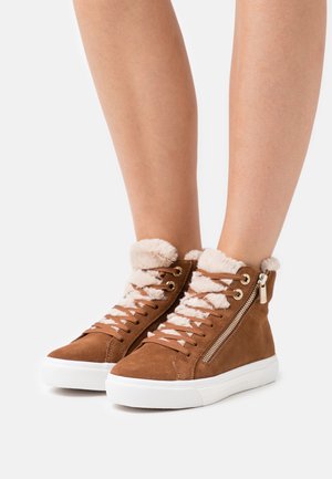 Baskets montantes marron avec doublure en fourrure blanche, lacets marron et fermetures éclair latérales portées sur des jambes nues sur fond blanc.