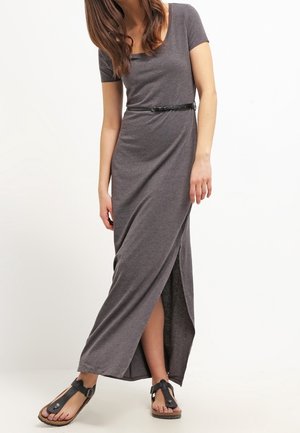 Femme portant une robe maxi grise à manches courtes avec une fente et une ceinture noire, associée à des sandales noires à boucle, debout devant un fond blanc.