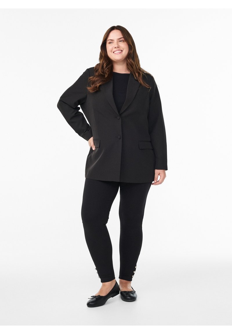 Mujer sonriendo con cabello largo y castaño, con una blazer negra holgada, leggings negros con botones cerca de los tobillos y bailarinas negras.