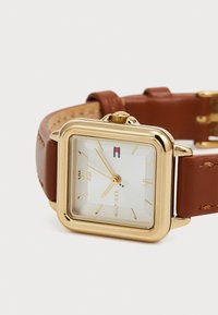 Reloj cuadrado dorado con una esfera blanca texturada, correa de cuero marrón y marcadores de números romanos. Cuenta con una corona para ajustes.