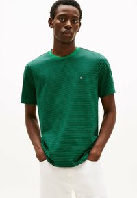 Grøn- og sortstribet kortærmet t-shirt med rund hals og logodetalje på brystet, parret med hvide bukser. Glat stofstruktur.