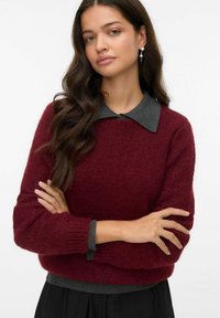 Burgunderfarbener Strickpullover mit einem grauen Kragen, strukturiertem Stoff, gerippten Bündchen und Saum, über einem schwarzen Rock getragen. Arme über Kreuz.