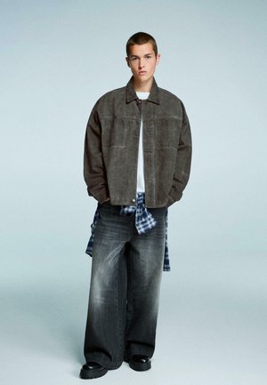 Giacca grigia in suede sovradimensionata su una camicia bianca, abbinata a jeans grigi sbiaditi a gamba larga e una camicia a quadri blu annodata in vita. Stivali neri.