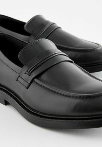 Svarta läderloafers med en slät finish, med en dekorativ rem och sömnadsdetaljer, placerade på en lågprofils gummisula.