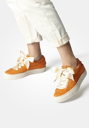 Sneaker in pelle arancione con suole in gomma bianca e nastri in satin di grandi dimensioni come lacci. Dettagli testurizzati sul tallone; visibili pantaloni chiari con risvolto.