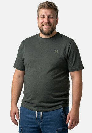 Uśmiechnięty mężczyzna z krótkimi włosami i brodą, ubrany w ciemnoszary, krótki rękaw koszuli oraz niebieskie spodnie z sznurkiem, stoi na tle jednolitego tła.