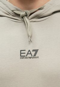 Világosszürke kapucnis pulóver húzózsinóros zárral, texturált anyagból, és egy feltűnő EA7 Emporio Armani logóval a mellkason.