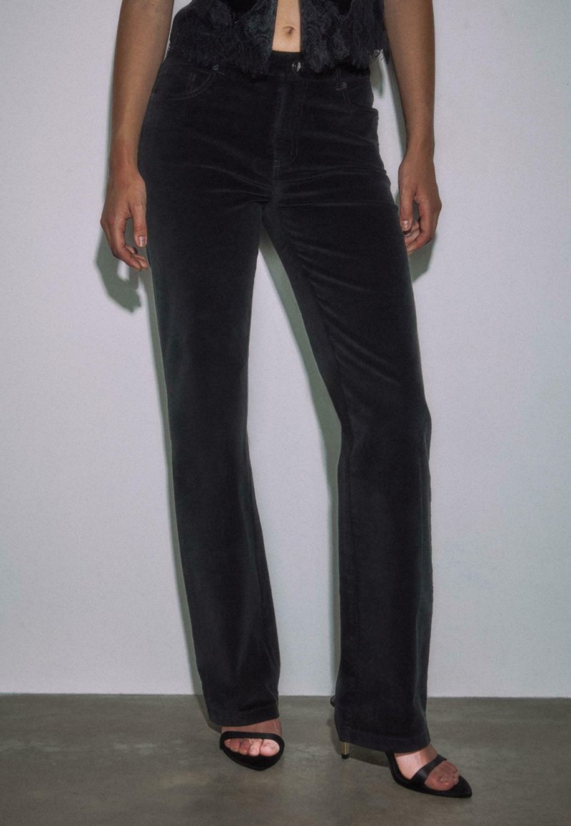 Kvinde iført tætsiddende sorte bukser og åbne sorte hæle, stående foran en ensfarvet lys væg.