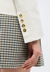 Λευκό μανίκι σακακιού με τέσσερα χρυσά κουμπιά ξεκουράζεται δίπλα σε χέρι που φοράει φούστα με ασπρόμαυρο σχέδιο ψαροκόκκαλο.