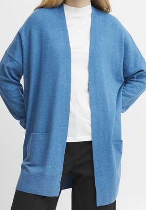 Kvinde iført en løs blå cardigan over en hvid top og sorte bukser, stående med hænderne bag ryggen mod en ensfarvet baggrund.