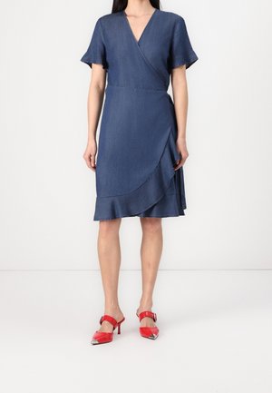 Femme portant une robe portefeuille bleue mi-longue à manches courtes avec un détail à volants, associée à des escarpins rouges pointus à bride-boucle.