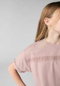 Lyserød kortærmet t-shirt med blondekant, der har teksten "94 ALLSA" på brystet. Blød tekstur med en afslappet pasform.