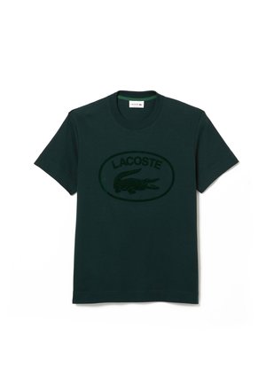 Dunkelgrünes Baumwoll-T-Shirt mit einem ovalen schwarzen Logo, das ein Krokodil-Graphic und das Wort "LACOSTE" in fetter, kontrastierender Schrift zeigt.