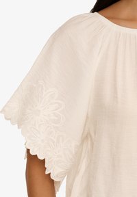 Blusa bianca con ampi maniche a scollop e ricami floreali. Tessuto leggero e testurizzato con dettaglio della scollatura arricciata.