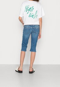 Donna con una T-shirt bianca con testo verde "High Tech", jeans corti blu e sandali bianchi senza lacci, in piedi con la schiena rivolta verso la fotocamera.