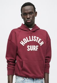Bordo duks s kapuljačom s ispisom "HOLLISTER SURF" u bijeloj boji, s prednjim džepom i rebrastim manšetama, izrađen od mekanog materijala.
