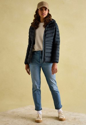 Musta pehmustettu vetoketjullinen takki, harmaa villapaita, vaaleansiniset farkut, vaaleanbeige kengät teksturoiduilla pohjilla ja vaaleanruskea lippalakki neutraalia taustaa vasten.