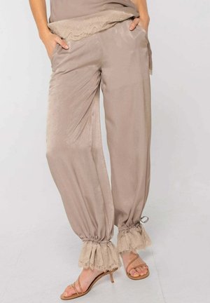Mujer con pantalones beige sueltos con puños de encaje fruncidos y sandalias de tacón con tiras color cuero, manos en los bolsillos, de pie sobre un suelo claro.