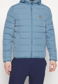Světle modrá péřová bunda s horizontálním prošíváním, předním zipem a kapucí. Má boční kapsy a malé žluté logo na hrudi.