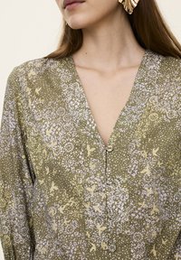 Grön blus med blommönster som har V-ringning och knappstängning. Tyget har en texturerad yta med små, fina blommönster.