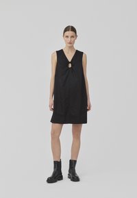 Robe noire sans manches avec un décolleté en V et une ouverture en trou de serrure, dotée d'un détail à nouer à la taille, en tissu lisse, longueur genou.