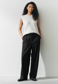 Lys grå ærmeløs sweater med ribbede kanter, parret med løstsiddende sorte bukser. Modelens hænder i lommerne, iført sorte sko.
