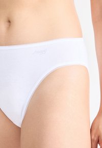Ingrandimento di una persona che indossa slip di cotone bianchi con bordi cuciti finemente e un sottile logo del marchio sulla cintura.