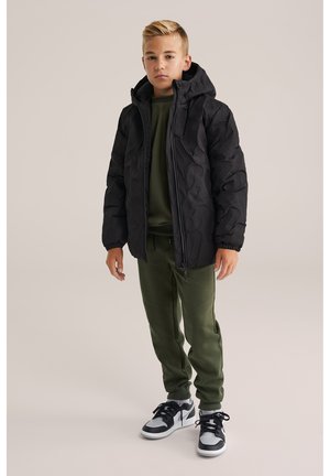 Garçon debout dans une veste à capuche noire à motifs, un pantalon de survêtement vert olive et des baskets noir et blanc sur fond uni.