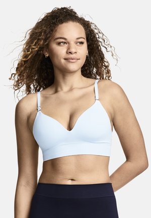 Frau mit lockigem Haar, die einen hellblauen Sport-BH und dunkle Trainingsleggings trägt, sieht mit neutralem Ausdruck leicht seitlich.