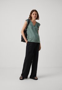 Groene V-hals top met textuur en gerimpelde mouwen, gecombineerd met brede zwarte broeken en zwarte schoenen. Zwarte tas over de arm.