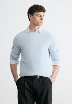 Άνδρας με κοντά σγουρά μαλλιά που φοράει ανοιχτό μπλε πουλόβερ με υφή πάνω από λευκό πουκάμισο με γιακά και μαύρο παντελόνι με λεπτές ρίγες, με τα χέρια στις τσέπες, κοιτάζοντας δεξιά.