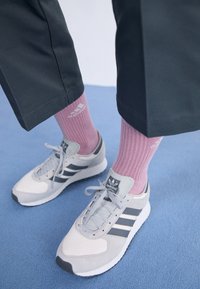 Gray Adidas sneakers with white soles worn with pink Adidas socks and cropped black pants on a blue carpeted floor.