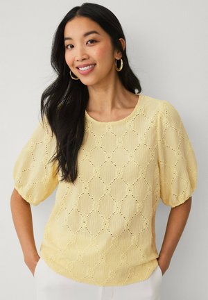 Młoda kobieta z długimi czarnymi włosami, nosząca złote kolczyki koła i teksturowaną jasnżółtą bluzkę z bufkami na rękawach, uśmiechająca się z rękami w kieszeniach.