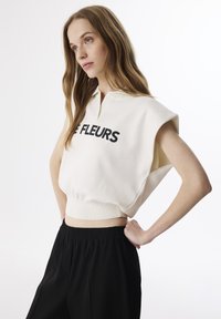 Felpa corta color crema con colletto e testo nero "LE FLEURS". Presenta ampi fori per le braccia e un orlo a coste, abbinata a pantaloni neri larghi.