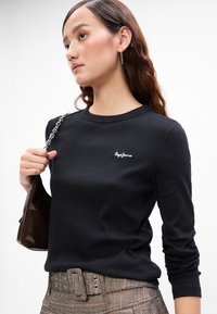 Fiatal nő hosszú hajjal, fekete Pepe Jeans hosszú ujjú pólót visel, kockás, magas derekú nadrágot, karikafülbevalót és egy barna táskát tart a kezében.