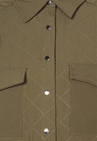 Camisa acolchoada verde-oliva com padrão de losangos, botões de pressão prateados e dois bolsos no peito com fechos de aba.