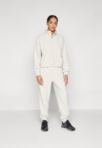 Beige fleece zip-up sweatshirt with a stand collar and matching joggers, featuring elastic cuffs and waistband, styled with black sneakers.