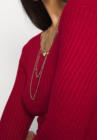 Pull en maille rouge côtelée avec un décolleté en V profond. Deux colliers en or superposés, l'un avec un pendentif en forme de cœur, l'autre une chaîne fine.