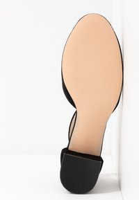 Vue unique d'une chaussure en suède noir avec une semelle beige lisse et un petit talon carré reposant contre un coin blanc.