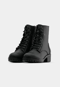 Bottines en cuir noir avec un laçage à l'avant, semelles texturées et talons blocs. Comprend une fermeture éclair sur le côté et des détails de couture renforcée.